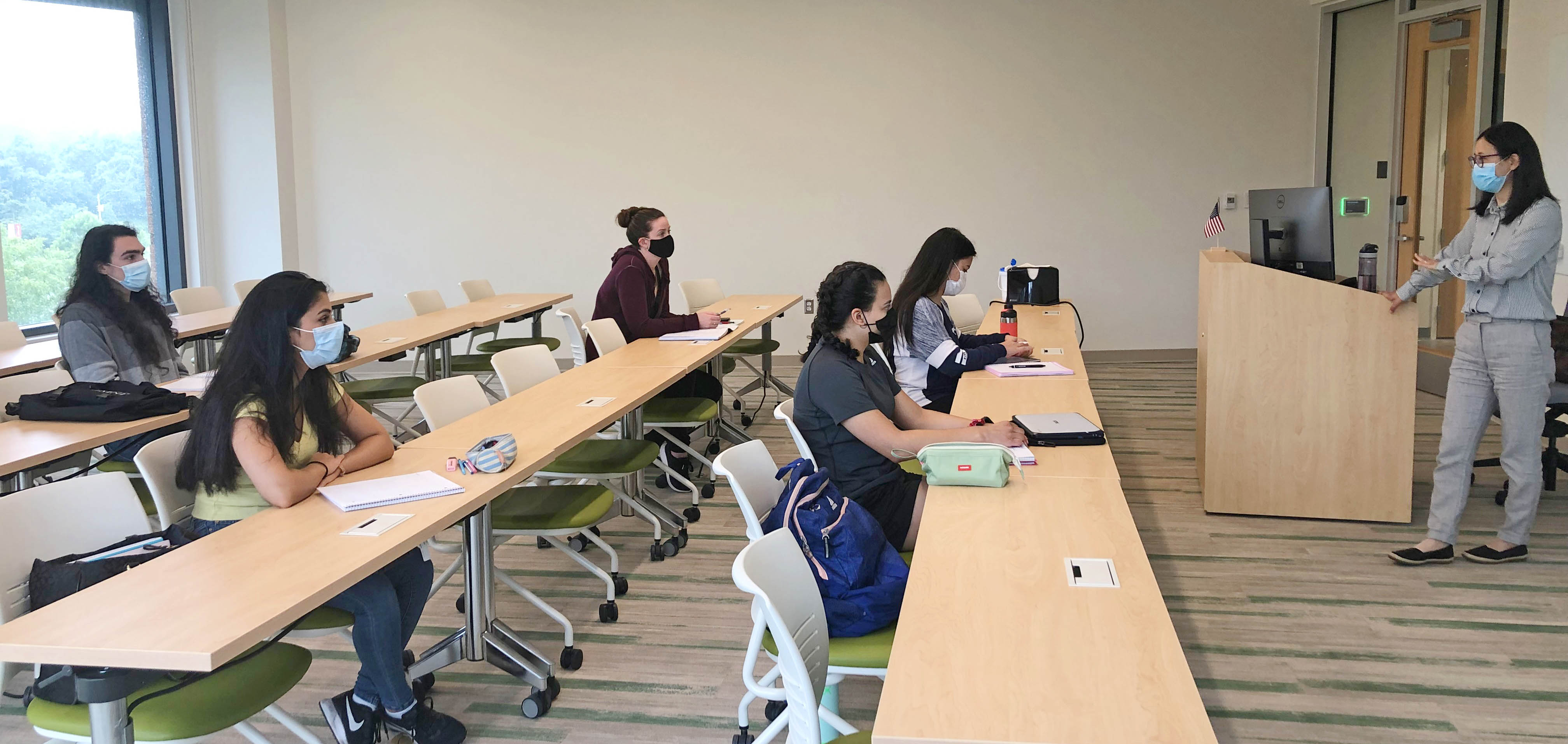 Dr. Lan Ma is pictured (right) at the front of a classroom teaching five students (left) who are taking notes. 